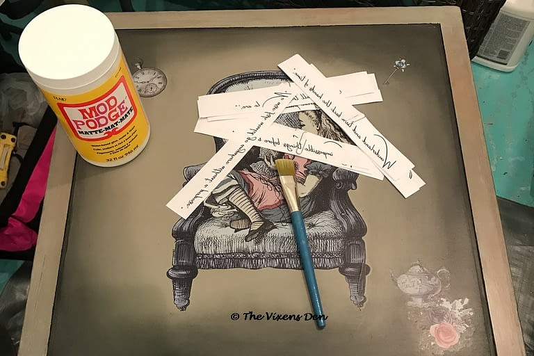 view of a table top with several Dixie Belle Alice in Wonderland transfers on the top of a square cherry side table with a jar of Mod Podge, strips of paper with printed quotes and a paintbrush