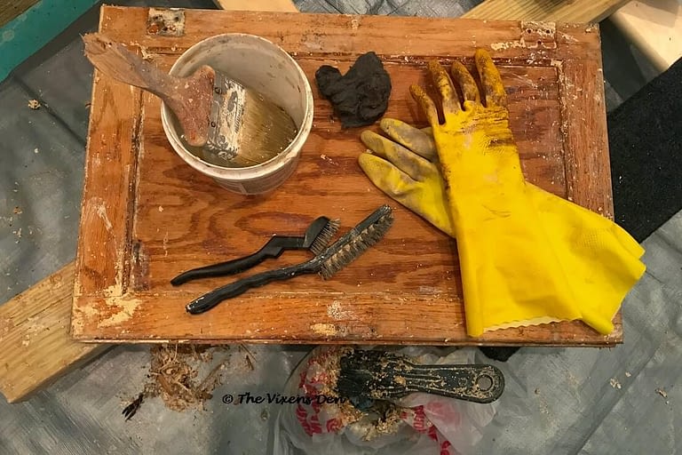 a cabinet door in the process of being stripped, with stripping supplies laid out: plastic container with a paintbrush, wire brushes, steel wool, rubber gloves, and a can lined with a plastic grocery bag with a plastic scraper balanced on top