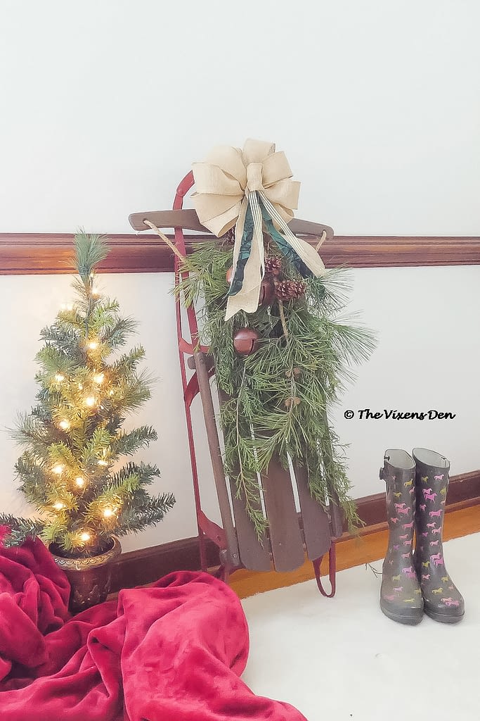 vintage sled staged with burlap bow, pine boughs, bells, boots, mini Christmas tree, and a red blanket