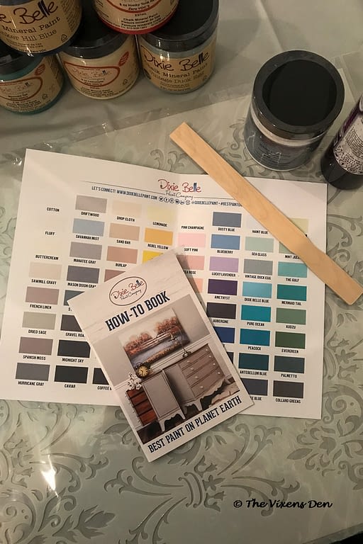 Dixie Belle paint swatch sheet with a paint stir and a How To Book, along with stacked paint and a jar of cleaner around the edges