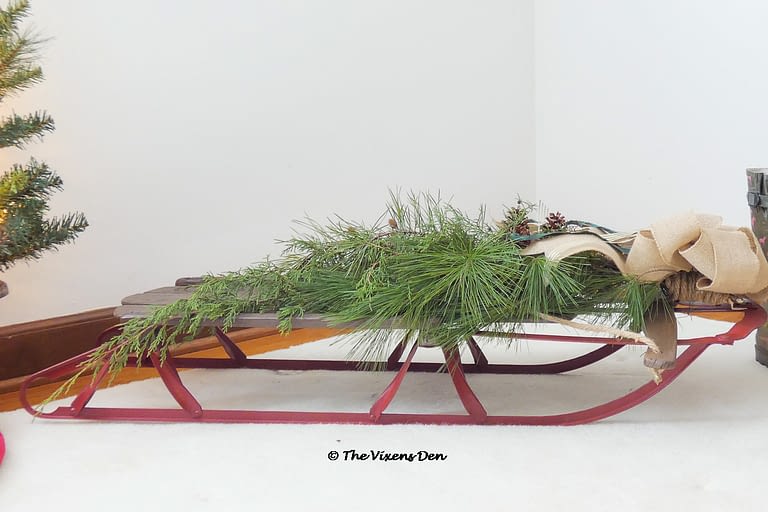 vintage sled staged with burlap bow, pine boughs, bells, boots, and a mini Christmas tree