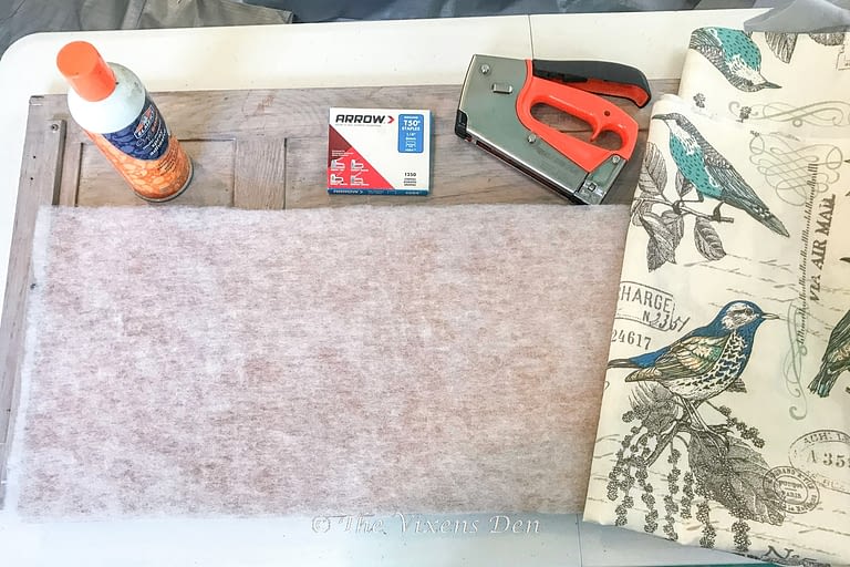 corkboard with luan backing and quilt batting applied and additional supplies ready to use: spray adhesive, staple gun, staples and fabric