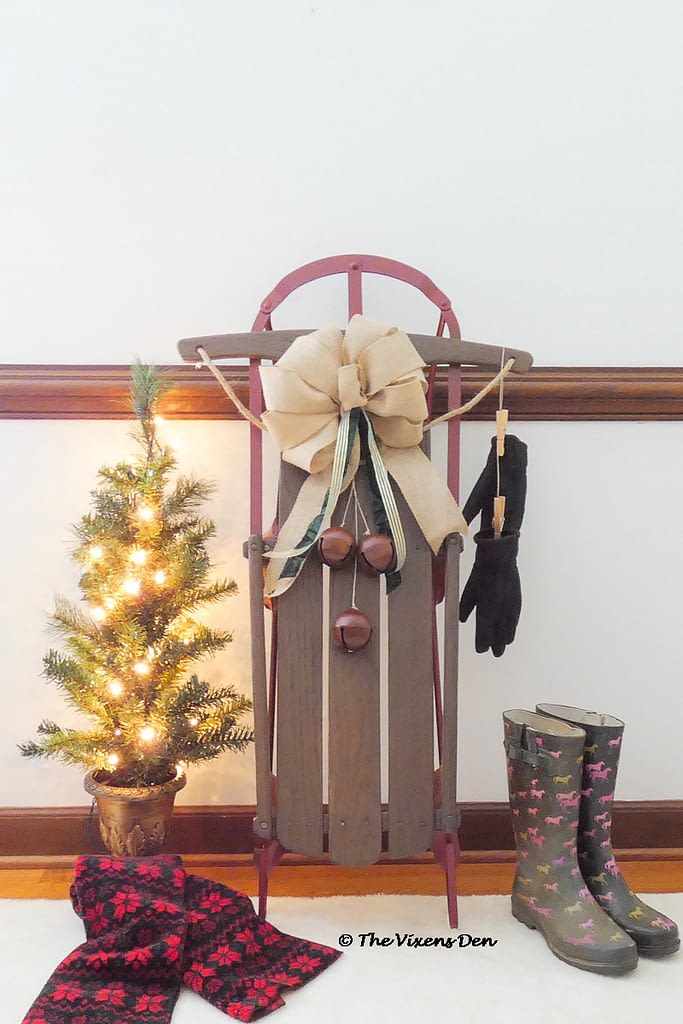 vintage sled staged with burlap bow, bells, boots, mini Christmas tree, black gloves, and a red and black scarf
